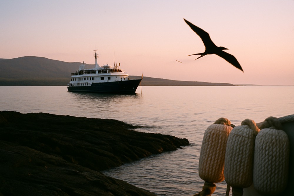 A small expedition ship anchored off a volcanic island shoreline at sunrise