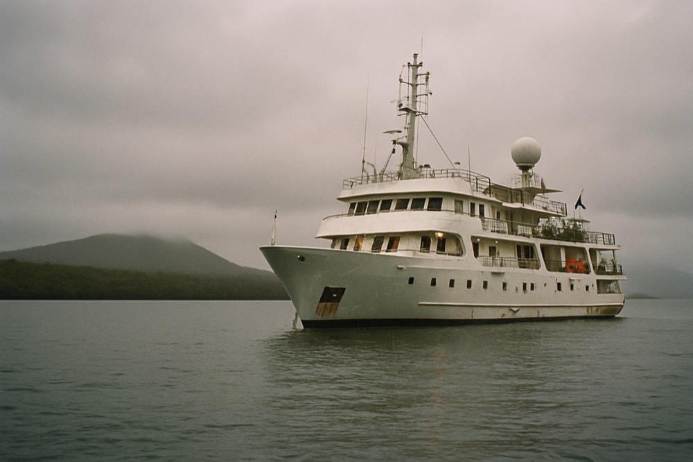An expedition cruise ship anchored near a volcanic island under low grey garua cloud
