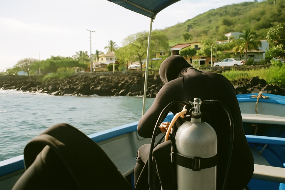 A diver preparing scuba equipment on a small boat beside a rocky shoreline