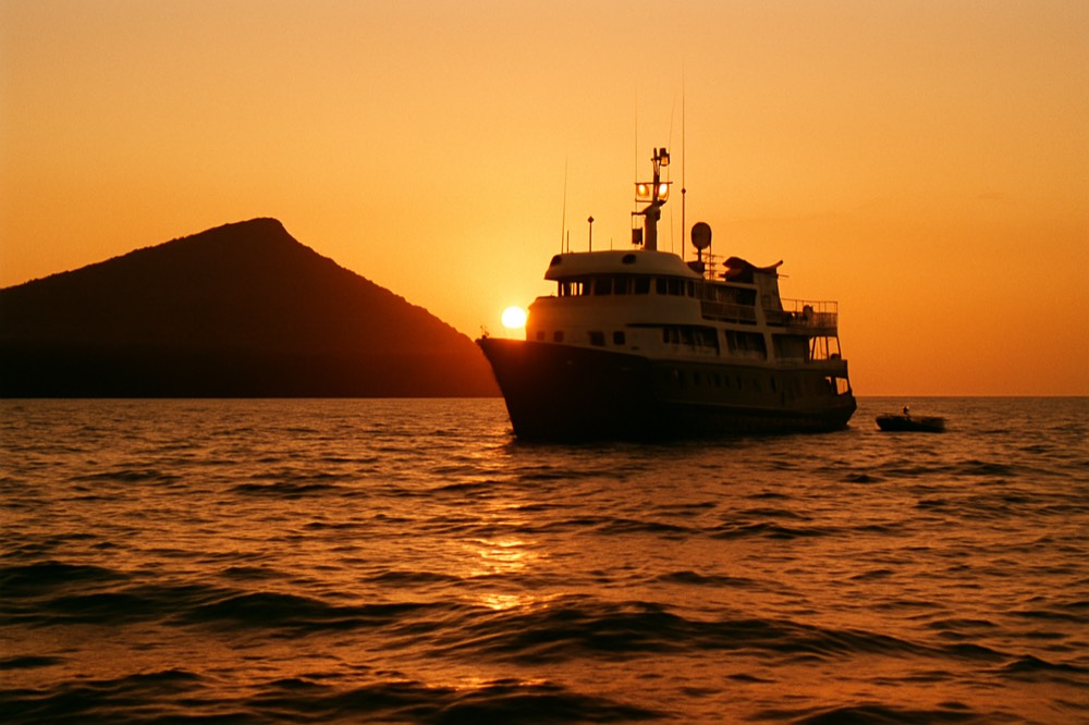 A small expedition vessel anchored off a volcanic Galapagos shoreline at sunset