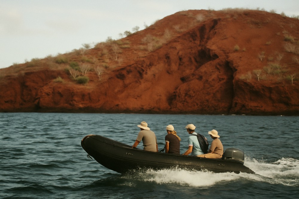 Travellers in a small inflatable boat crossing open water toward a volcanic shore